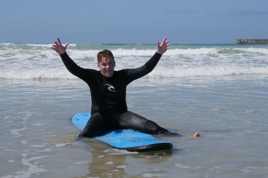 school teen sits in shallows of ocean on surf board with his arms flung in the air