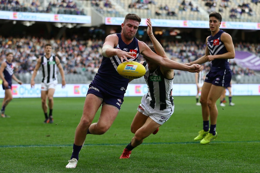 AFL footballer eyes the ball as he attempts to kick it upfield while a defender tries to smother it.