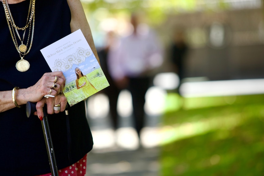A woman holds a walking stick and a funeral booklet with a young woman on the cover. 