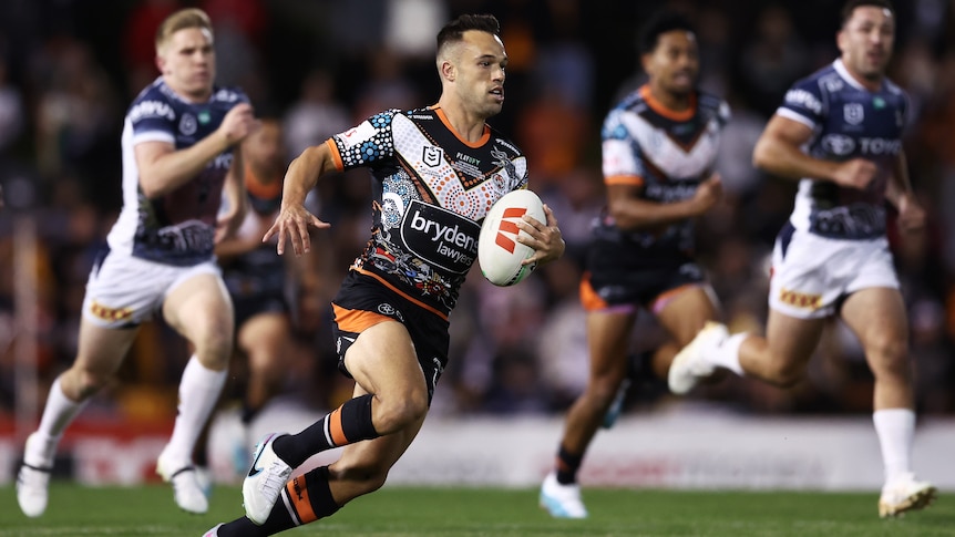 A Wests Tigers NRL player holds the ball with his left hand as he makes a break against North Queensland.