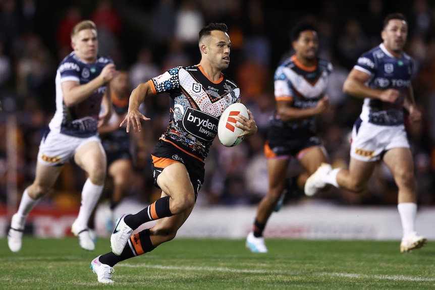 A Wests Tigers NRL player holds the ball with his left hand as he makes a break against North Queensland.