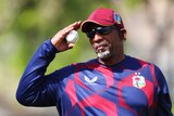 A West Indies cricket coach stands wearing dark glasses, holding a ball in one hand as he watches the play.