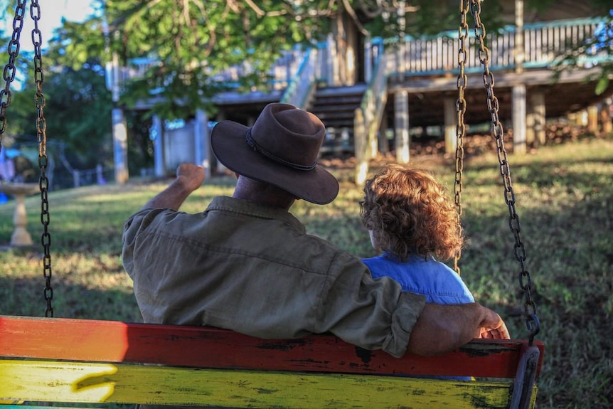 A still from the film Don't Tell: two characters sit on an outdoor swing, facing away form the camera.