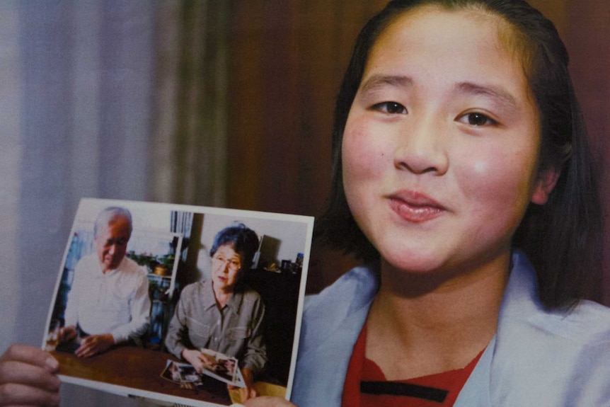 A young Japanese girl holds up a photo of an elderly Japanese couple