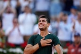 A young tennis star stands on court with a beaming smile on his face and his hand on his heart.