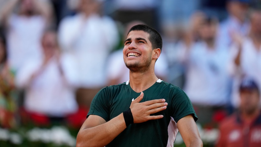 A young tennis star stands on court with a beaming smile on his face and his hand on his heart.