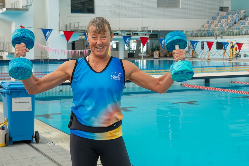 Woman with grey hair in ponytail stands to the left of a swimming pool holding up two foam dumbells.