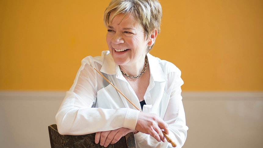 Conductor Marin Alsop smiles looking away from the camera. She sits on a chair holding a baton, in front of a yellow wall.