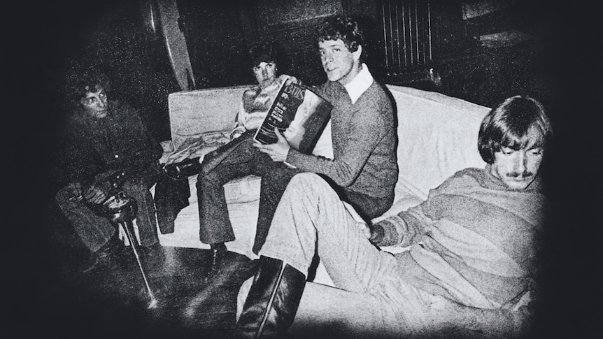 Grainy, black and white photo of the Velvet Underground sitting on a couch. Lou Reed holds a magazine.