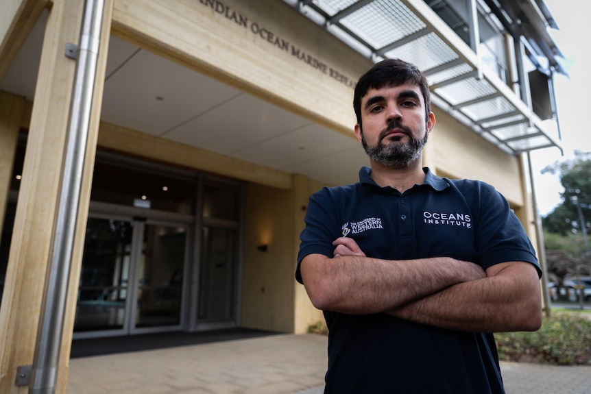 A man in a blue polo with a logo saying Oceans Institute, standing in front of a building with his arms folded