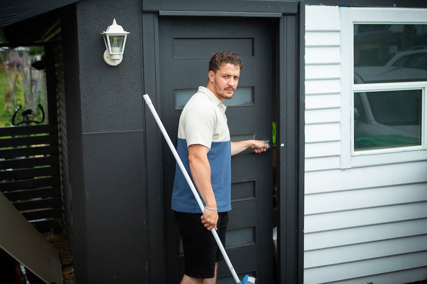 A man with a broom is about to enter a shed, looks back at the camera.