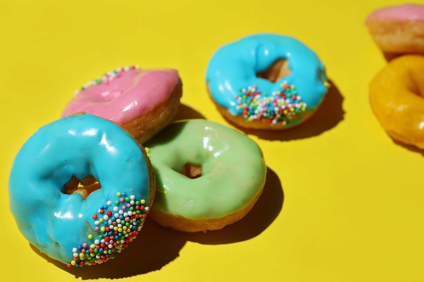 A stack of brightly coloured ice doughnuts with hundreds and thousands sit on a coloured background, a nostalgic sugar fix.