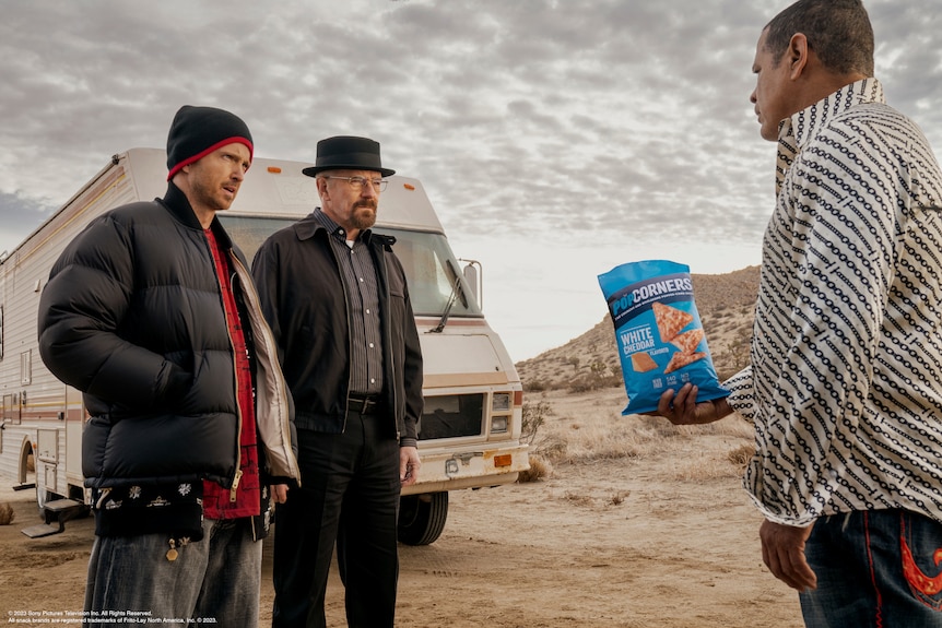 Bryan Cranston and Aaron paul stand in front of Raymond Cruz who holds out a chip packet.
