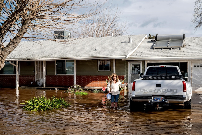 A woman up to her knees in flood water next to a car outside a house
