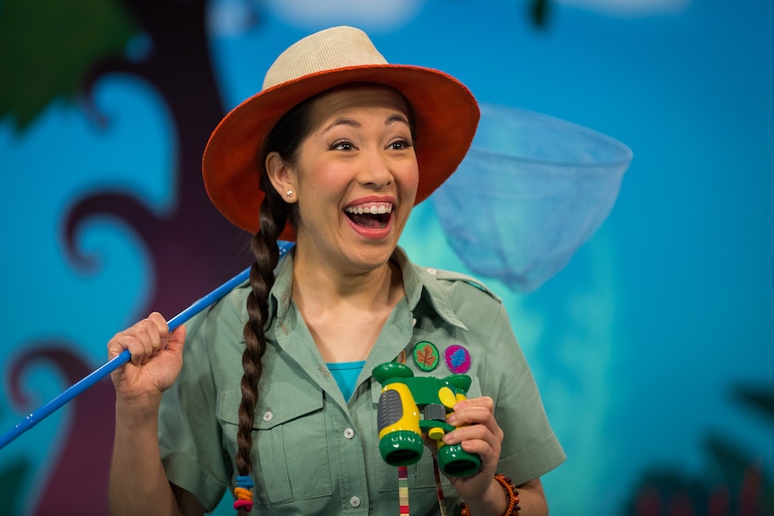 An Asian Australian woman wears an explorer's outfit and broad-brim hat, looking excitedly off-camera while holding binoculars