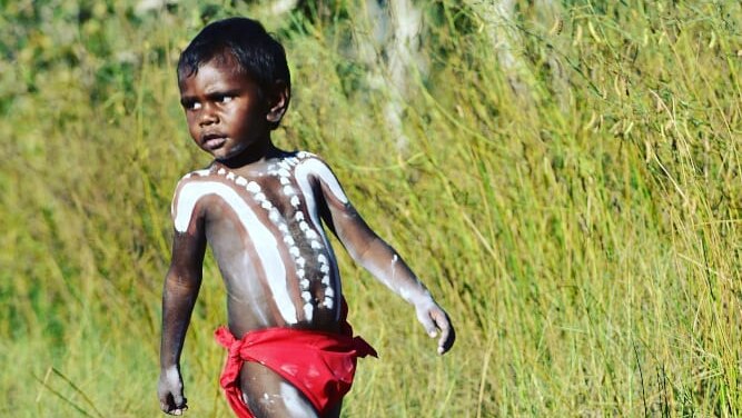 A young boy in traditional Indigenous paint and clothing walks along a dirt track