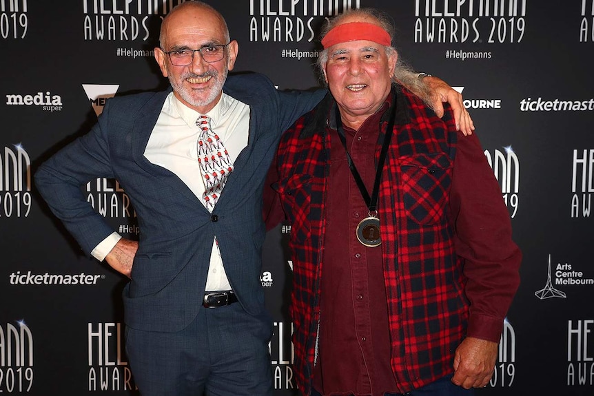 The two musicians stand with their arms around each other's shoulders in front of a Helpmann's media wall.