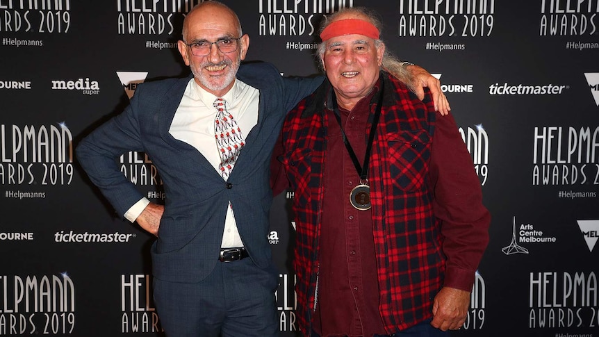 The two musicians stand with their arms around each other's shoulders in front of a Helpmann's media wall.