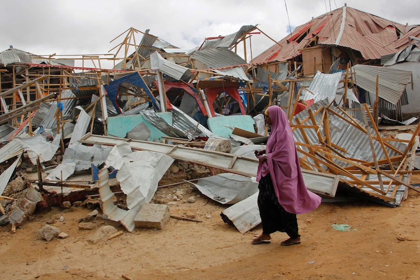 A woman in a pink headdress walks past broken buildings of timber and corrugated iron.