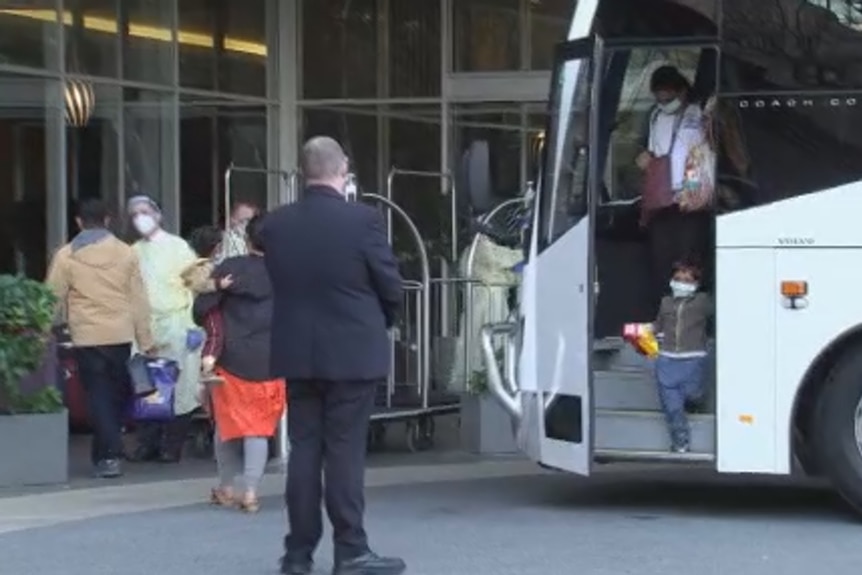 A child and a woman get off a bus as a man in a suit directs them towards the hotel entrance among other travellers.