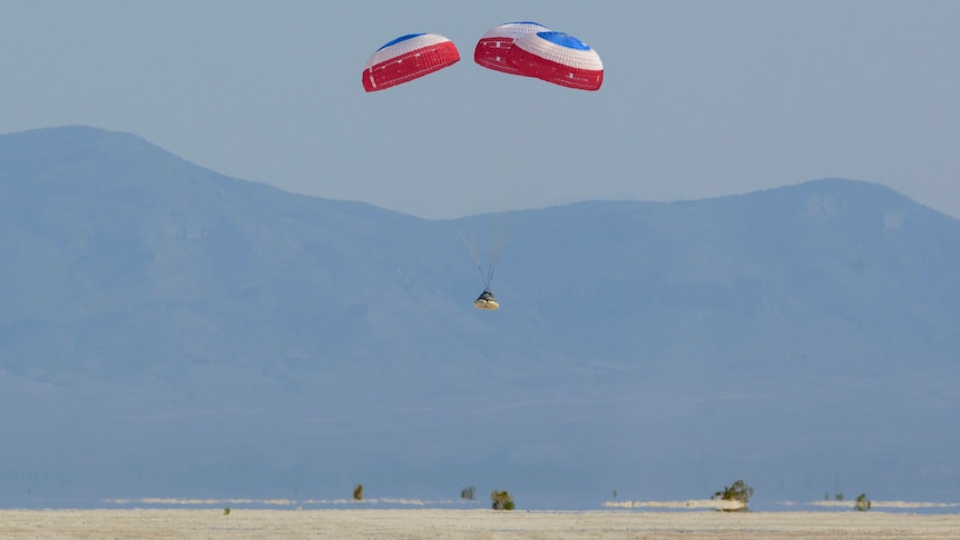 A small capsule is suspended under three red white and blue parachutes in the desert