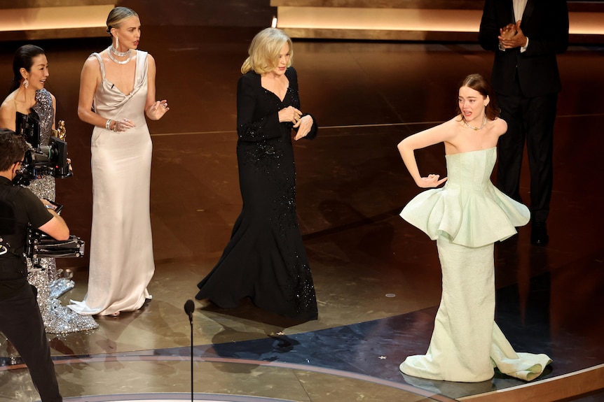 Emma stone pointing towards the back of her dress on stage surrounded by four other women. 
