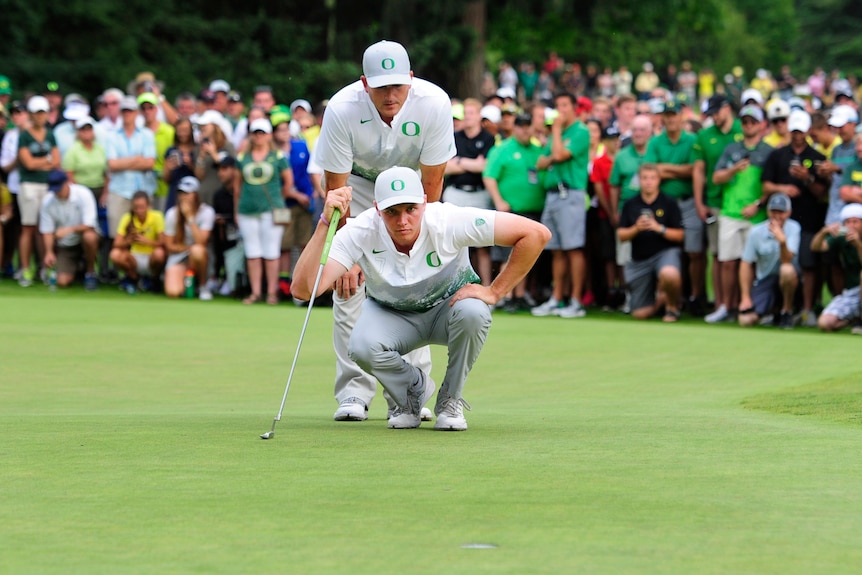 A US collegiate golfer crouches to look at putt as his coach stands behind him, both men wearing caps and shirts with "O" logos.