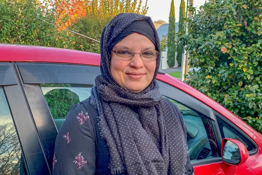 A woman in a headscarf stands against a red car with a slight smile on her face