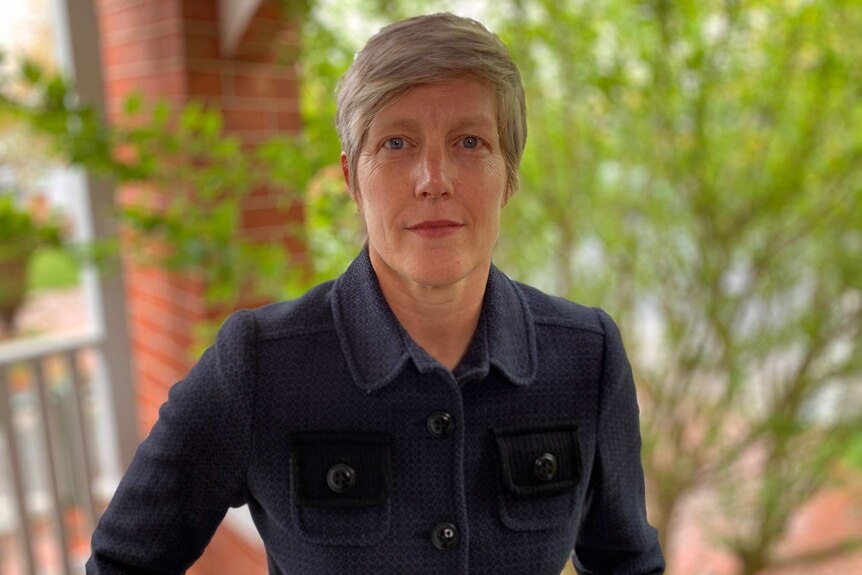 A woman with very short, fair hair and wearing a navy blue jacket looks into the camera.