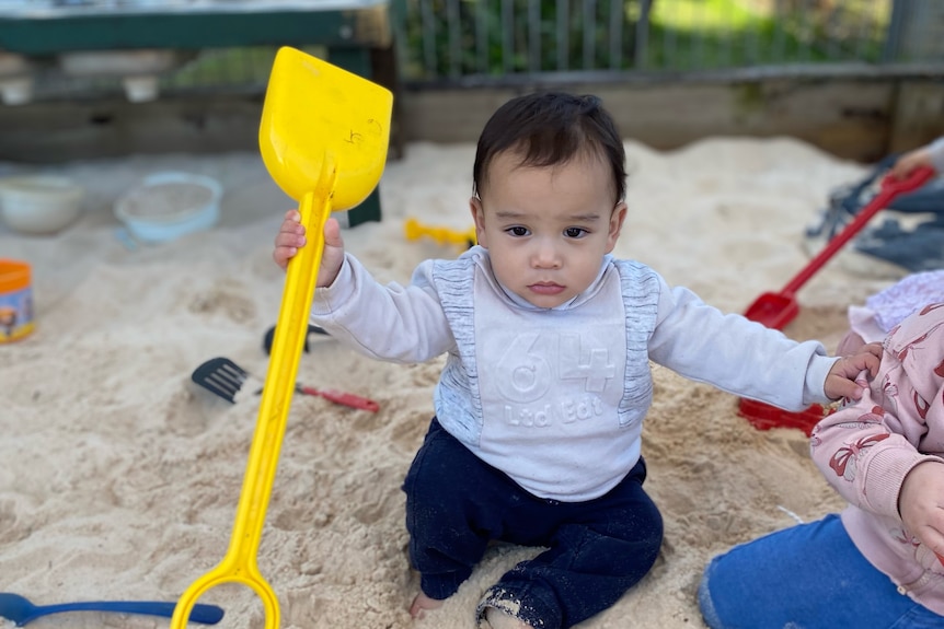 A toddler holding a shovel in a sandpit.