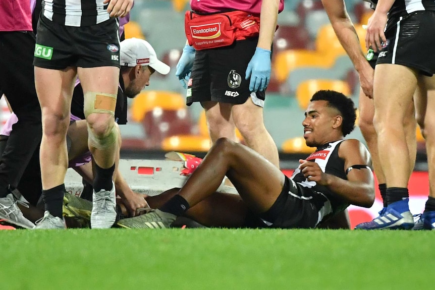 An AFL player lies on ground grimacing as trainers look at his leg injury with a stretcher nearby.