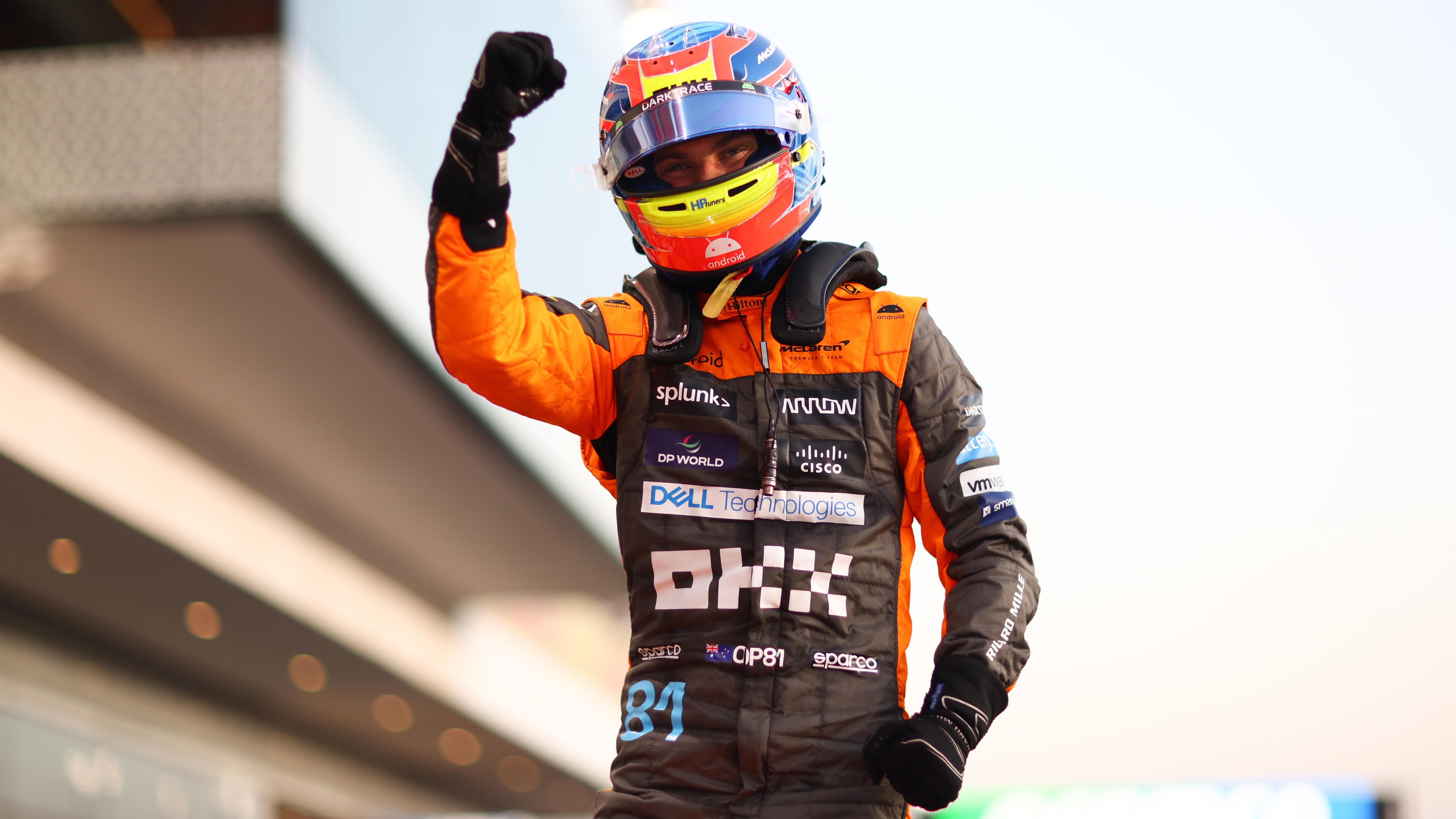 An F1 driver in an orange race suit, multi-colour helmet, standing on his car, raising his first in celebration