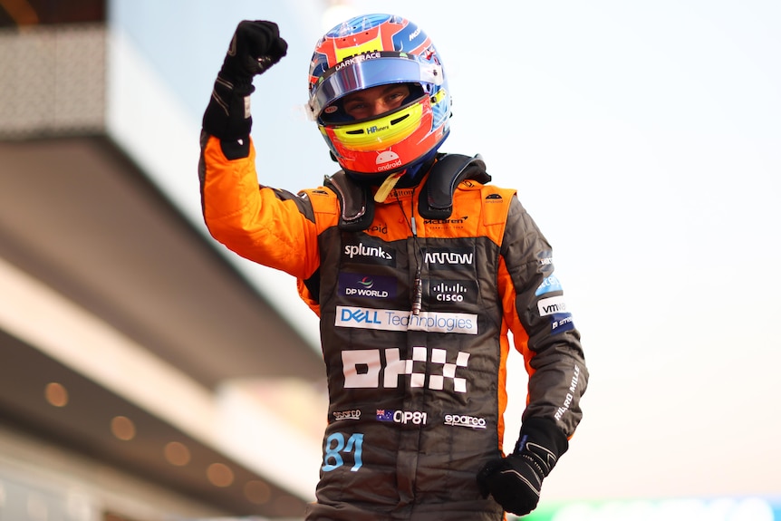 An F1 driver in an orange race suit, multi-colour helmet, standing on his car, raising his first in celebration