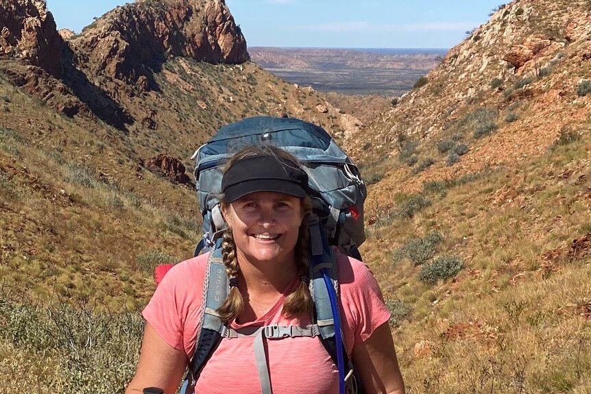 A smiling woman with a cap and a big backpack with mountains and desert in the background.