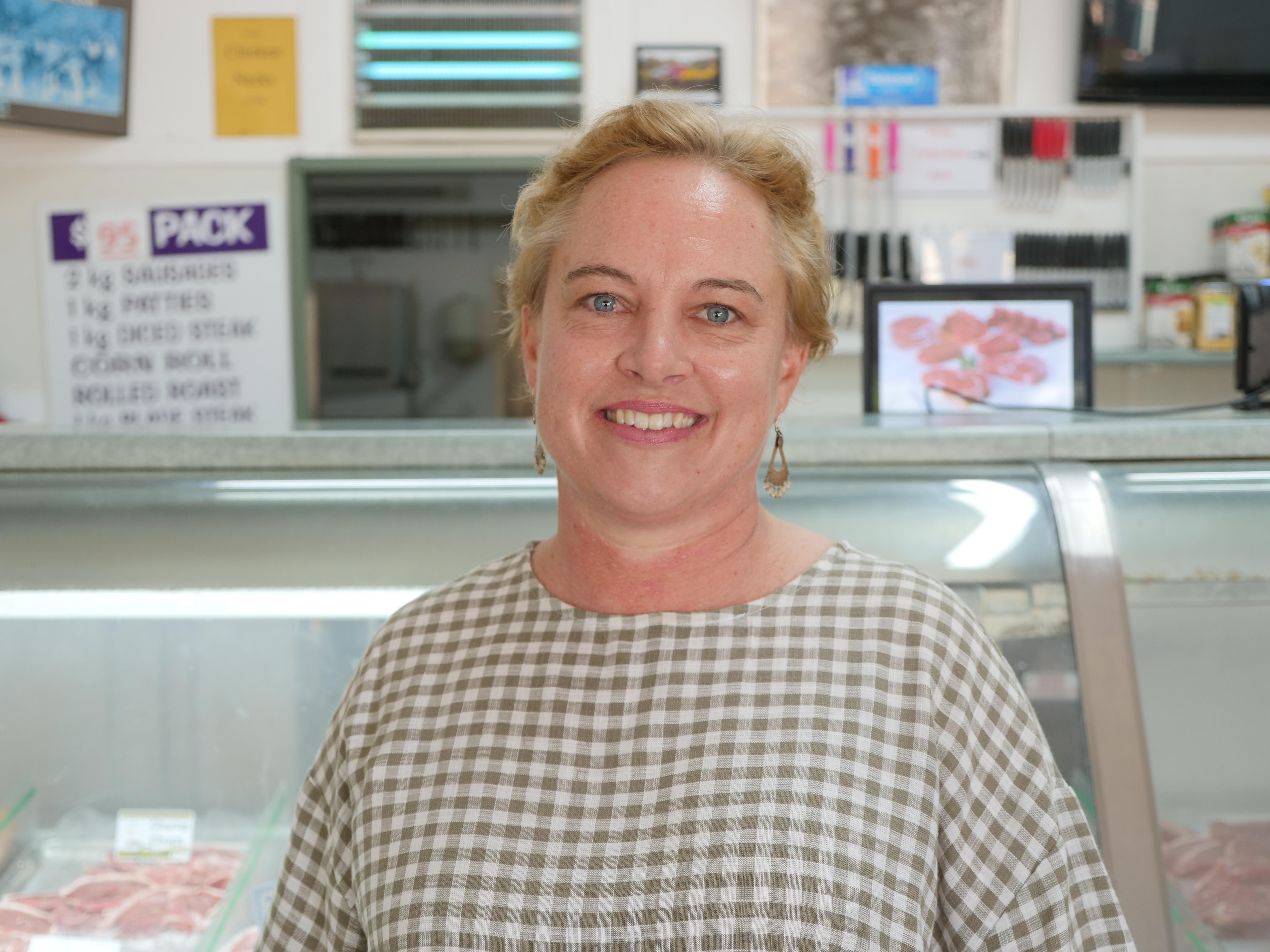 কসাইয়ের দোকানে মাংসের কাউন্টারের সামনে দাঁড়িয়ে চেক শার্ট পরা একজন মহিলা ক্যামেরার দিকে তাকিয়ে হাসছেন।