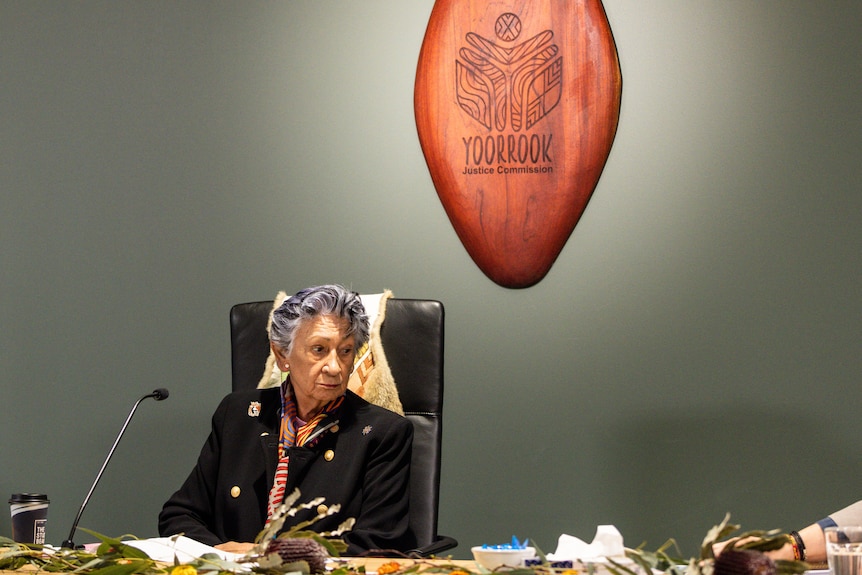 Eleanor Bourke wearing a black blazer and sitting in a chair with a possum cloak draped on it, looking pensively to the side.