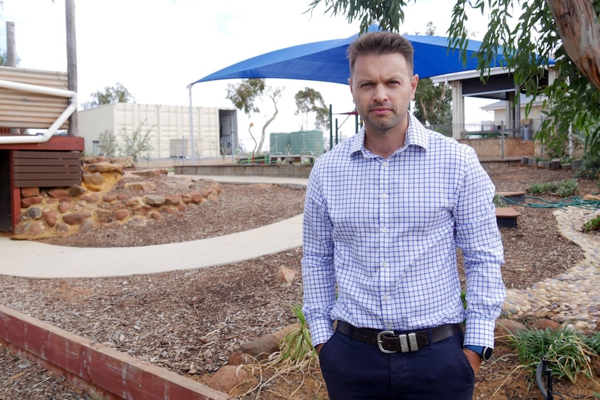 Man with checked blue shirt standing in front of a covered play area and neat landscaping at a school. 