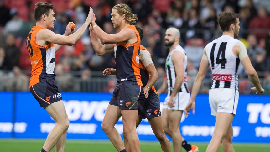 Two AFL players high-five in celebration after a goal, as their opponents walk past.