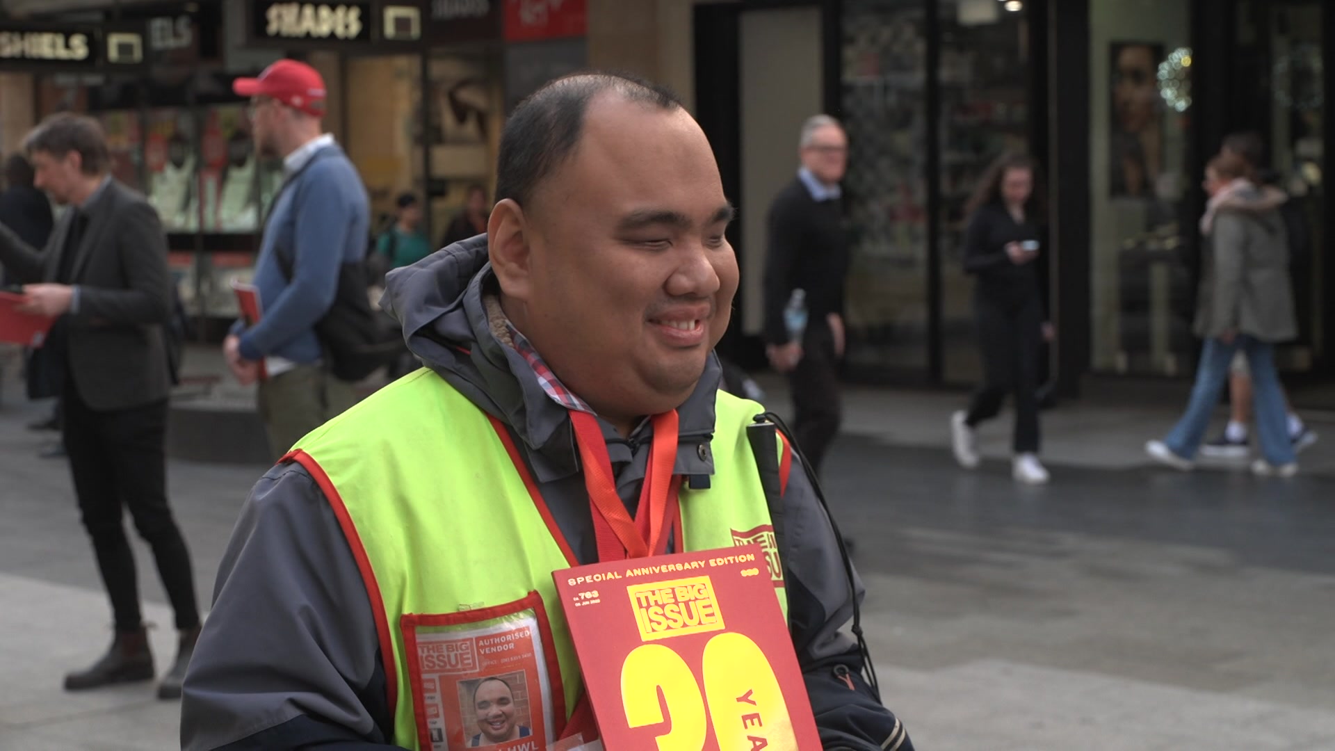 A man with his eyes closed wearing a red lanyard and fluorescent vest holding a red magazine smiles