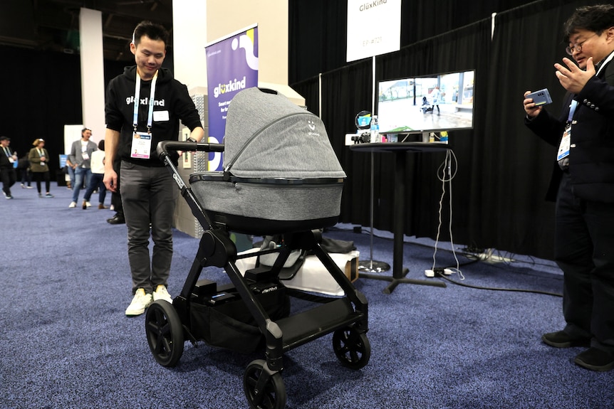 A medium shot of a man pushing an electric stroller indoors, as a man filming on a phone nearby watches on