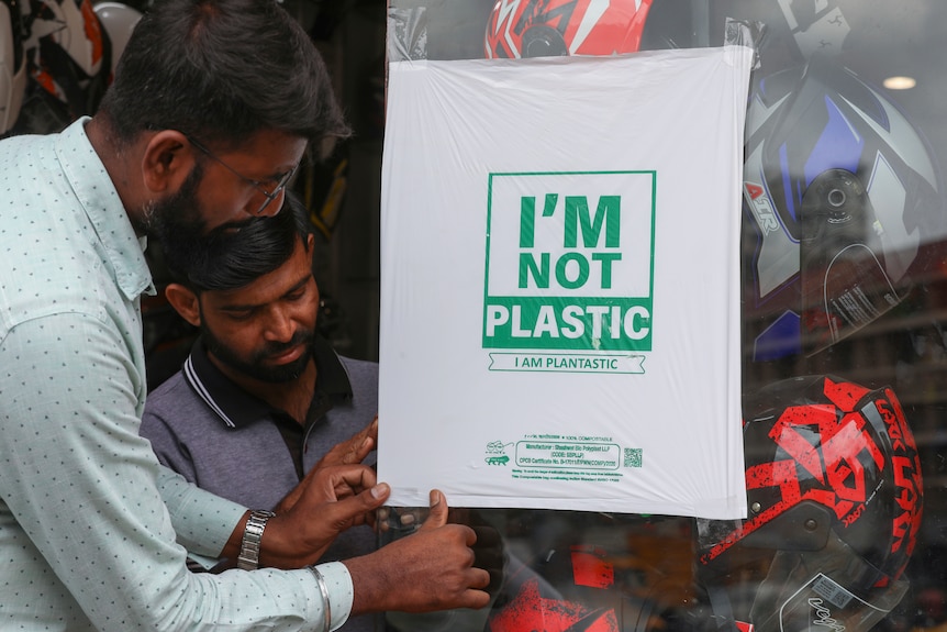 Two men inspect a stack of clear material designed to replaces single use plastic at a motorbike helmet shop.