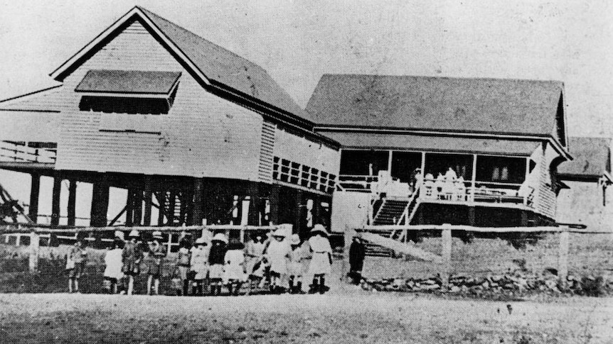 A black and white 19th century image of the school with children lining up for class.