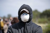 A person with the hood of their jacket raised over a cap, also wearing a mask, with only closed eyes visible.