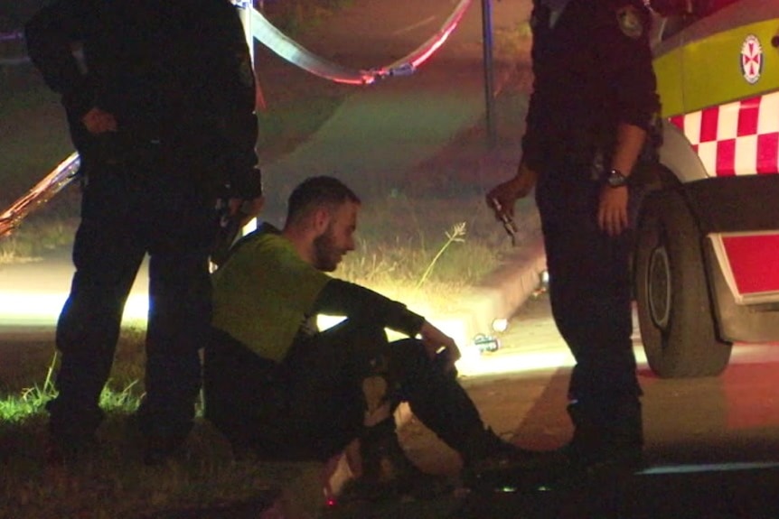 A man sits in a gutter with a police officer standing on either side of him and a parked ambulance.