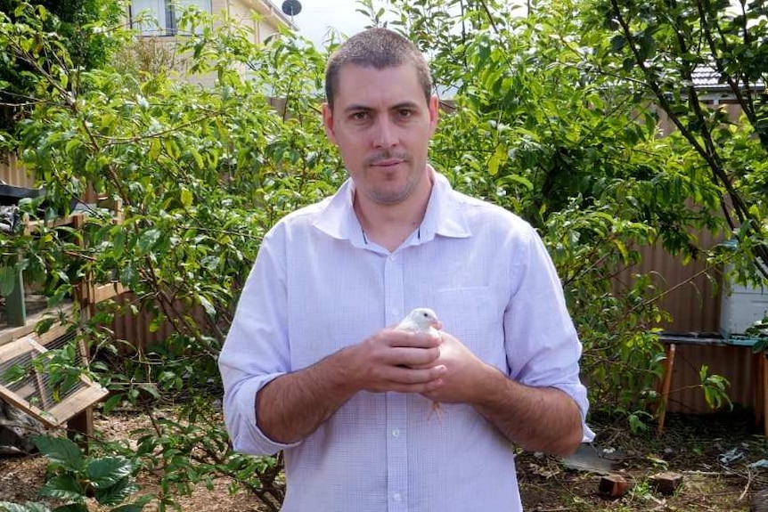 A man with short cropped hair stands in his backyard holding a small white bird.