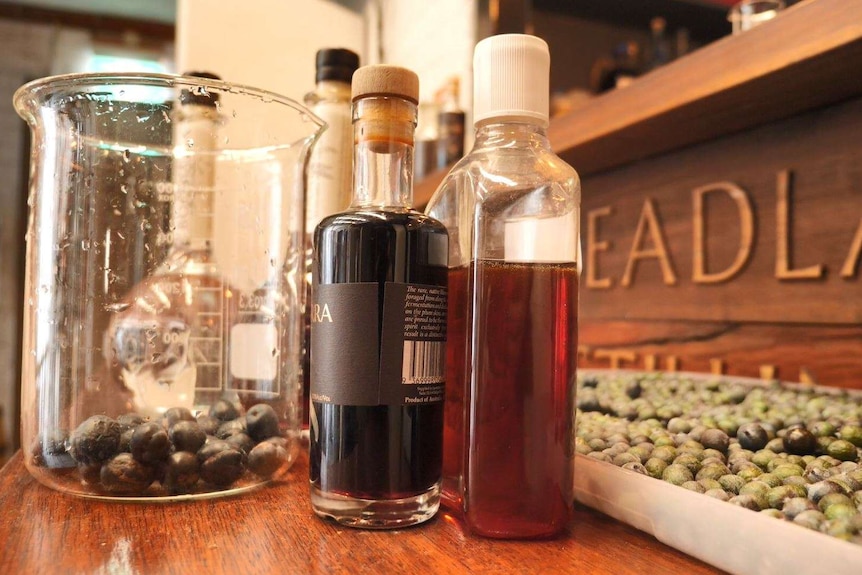 Bottles of spirits and honey on a table next to a beaker with Illawarra plums in it and seeds in the background.