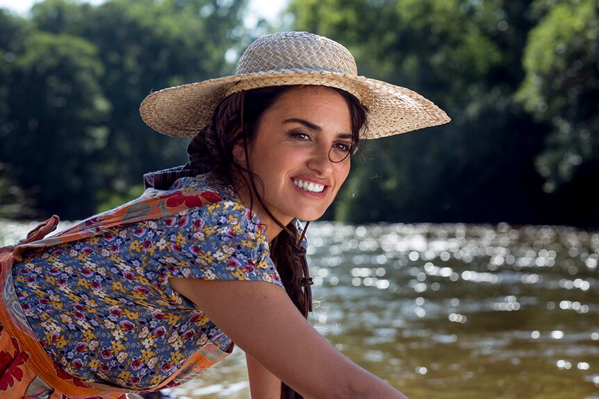 Penélope Cruz wears straw hat and smiles while kneeling with outstretched arm near bushy stream on sunny day.