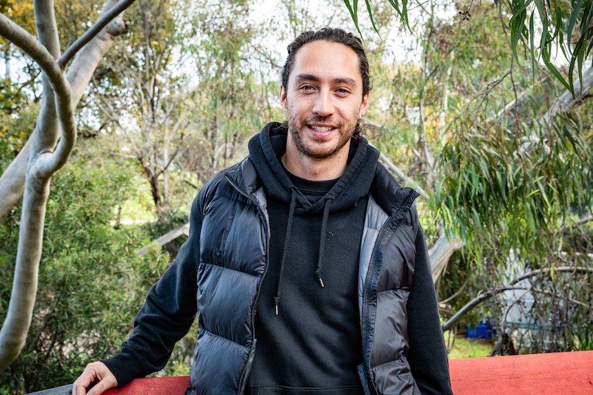 A young man with olive skin and a black puffer jacked standing in a garden and smiling