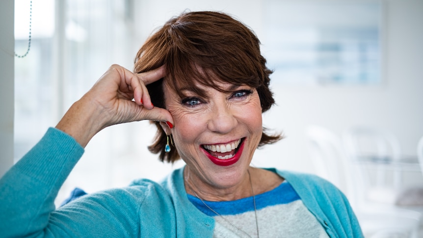 A woman with short brown hair wearing bright blue cardigan leans with hand on her forehead, big smile