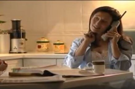 A woman sits at a kitchen table holding a cordless phone